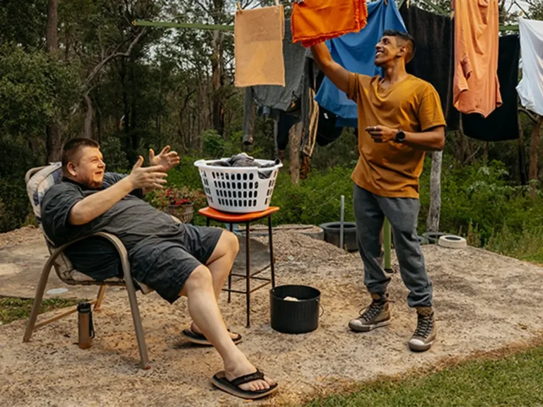 Support worker hangs washing on Hills Hoist clothesline while their client with disability talks.