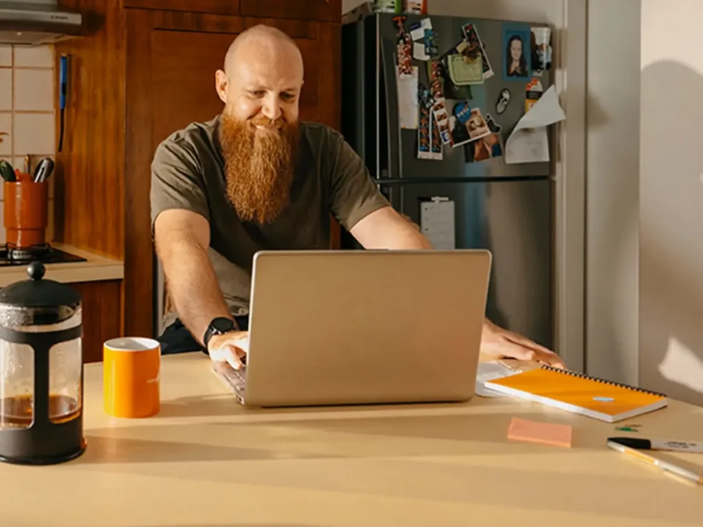 Support worker sits at kitchen bench with laptop and coffee to manage clients on the Mable platform.