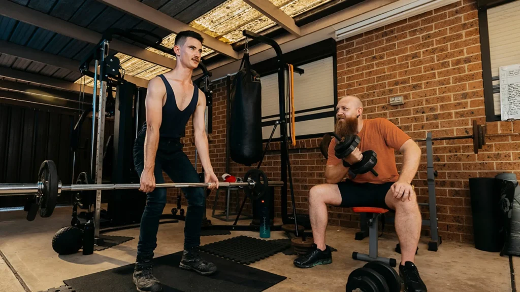 Support worker and young adult with autism and Tourette syndrome lift weights together in home gym.