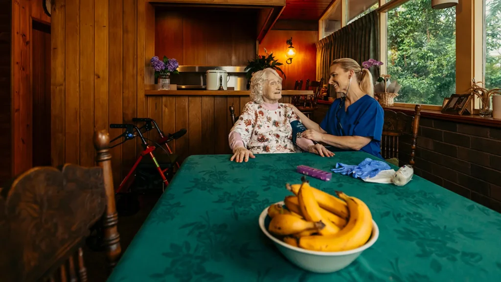 Registered Nurse takes a blood pressure reading for their older client at their dining table. 