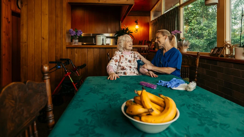 Registered Nurse takes a blood pressure reading for their older client at their dining table. 