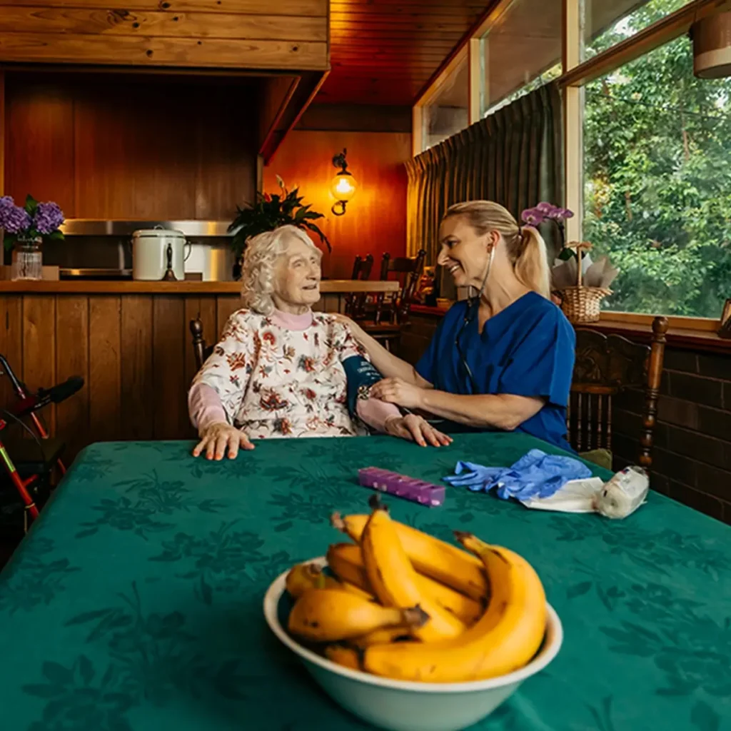 Registered Nurse takes a blood pressure reading for their older client at their dining table. 