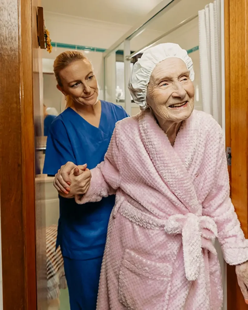 Registered Nurse in scrubs helps older client in pink bathrobe walk out of their bathroom.