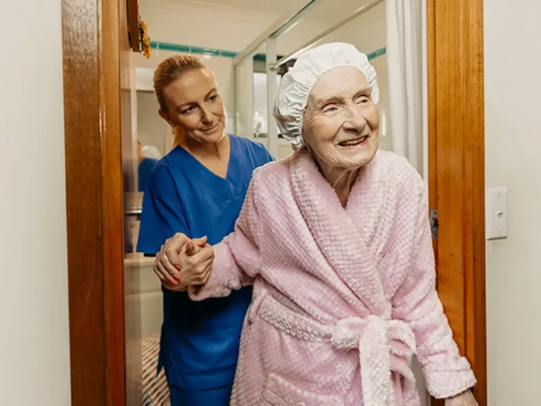 Registered Nurse in scrubs helps older client in pink bathrobe walk out of their bathroom.