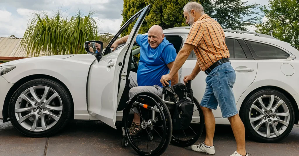 Older support worker holds wheelchair at car door for client who is blind and paraplegic.