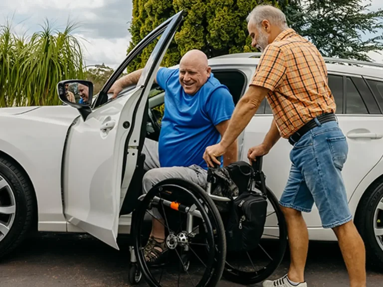 Older support worker holds wheelchair at car door for client who is blind and paraplegic.