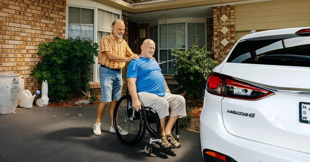 Older support worker helps client who is blind and paraplegic wheel across the driveway to a car.