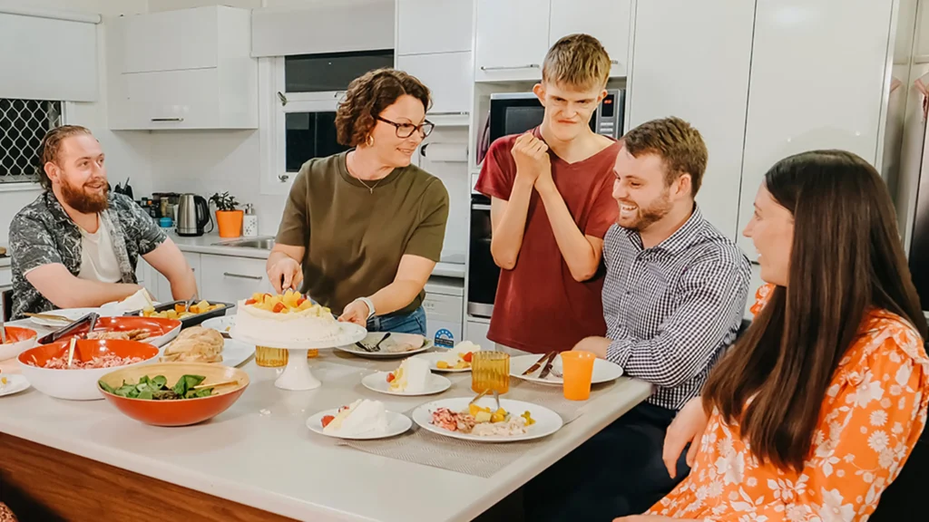 Adult with disability grins at pavlova on the dinner table while support team and friends chat.