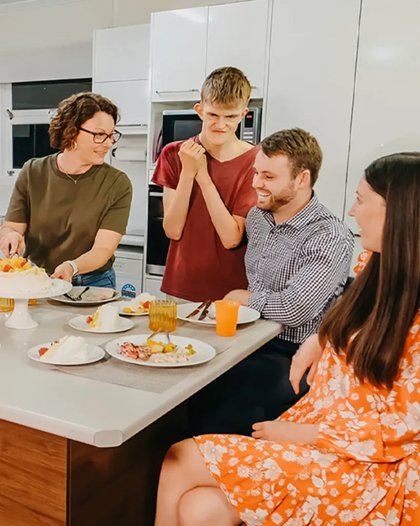 Adult with disability grins at pavlova on the dinner table while support team and friends chat.