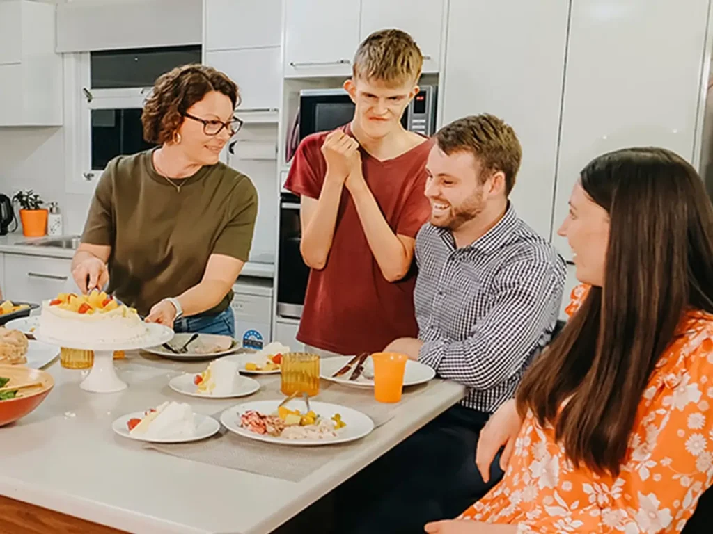 Adult with disability grins at pavlova on the dinner table while support team and friends chat.