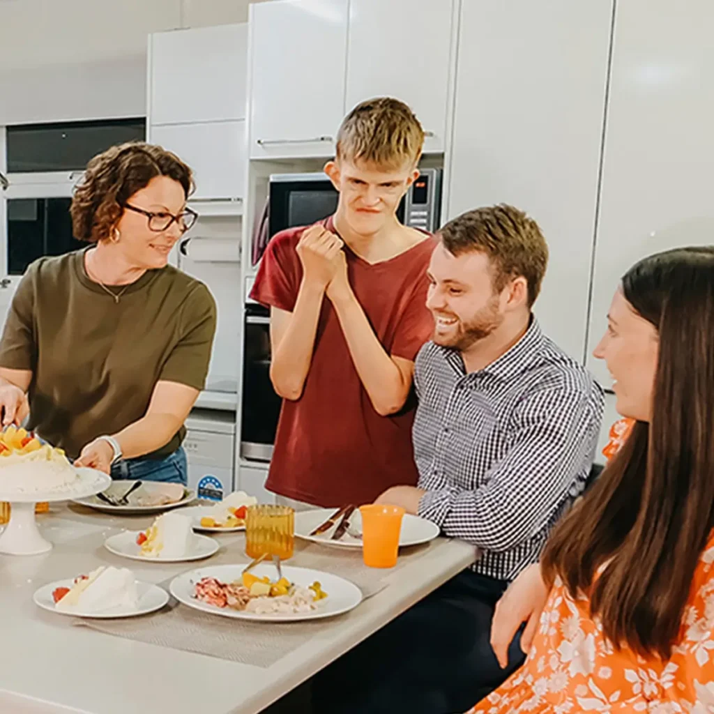 Adult with disability grins at pavlova on the dinner table while support team and friends chat.