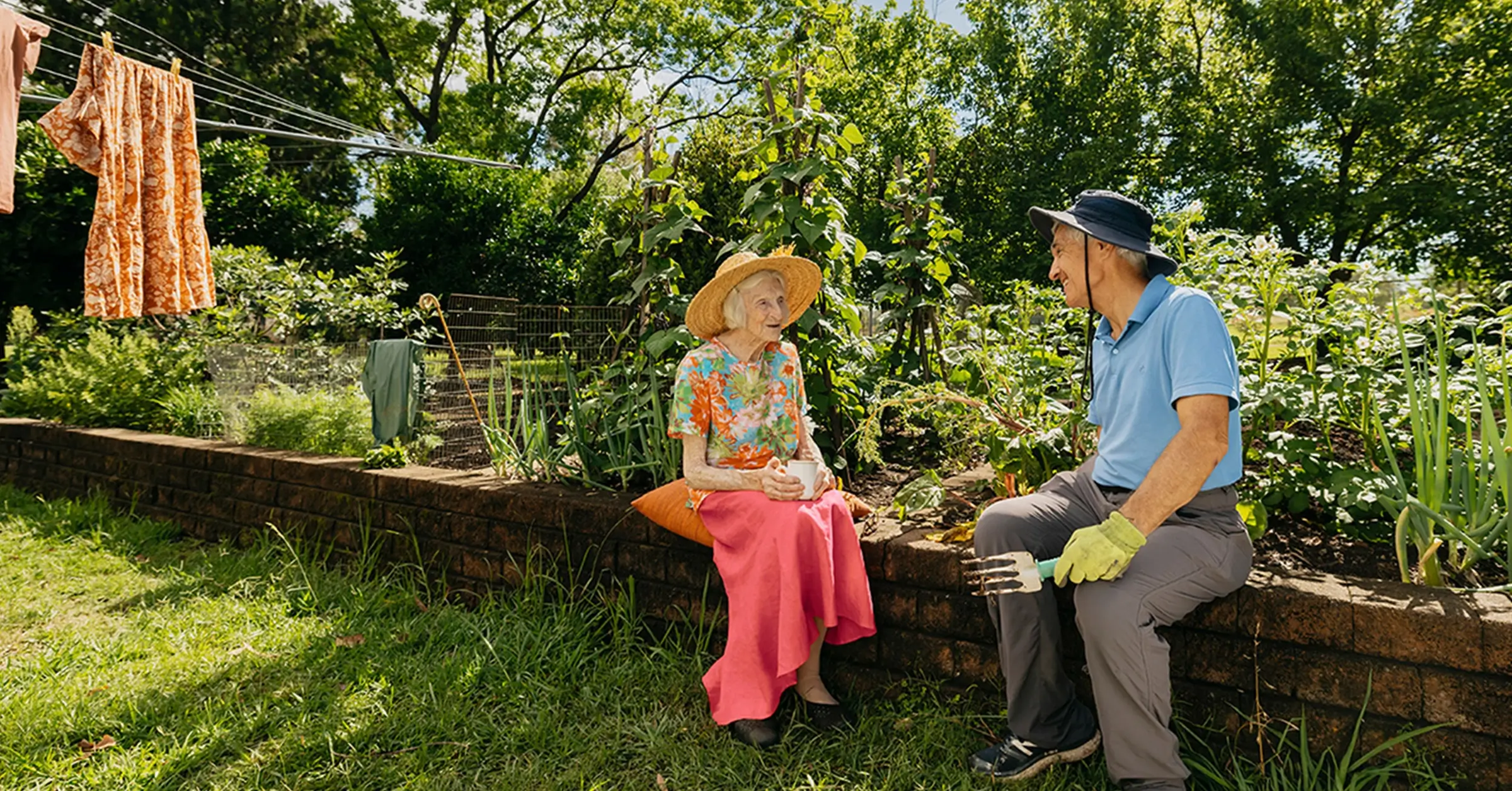 Support worker prunes plant in garden while chatting to their older client sitting on garden wall.