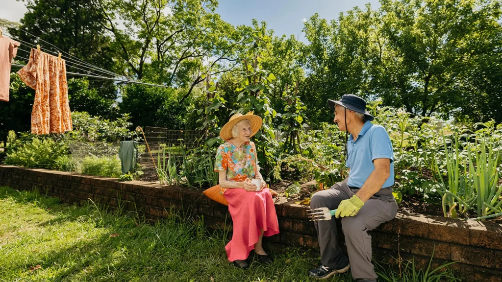 Support worker prunes plant in garden while chatting to their older client sitting on garden wall.