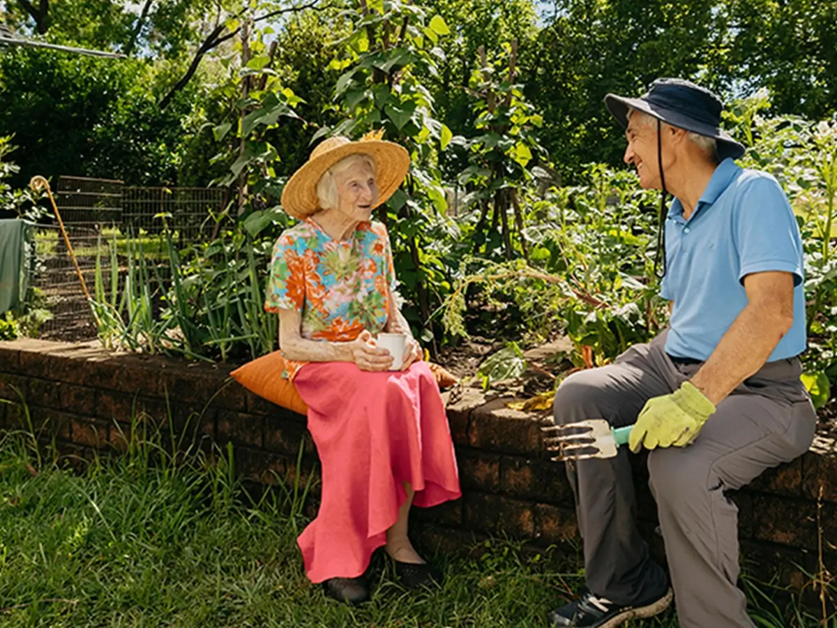 Support worker prunes plant in garden while chatting to their older client sitting on garden wall.