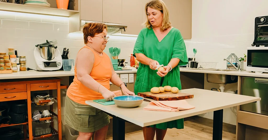 Person with disability prepares food at a low table in their accessible kitchen with support worker.