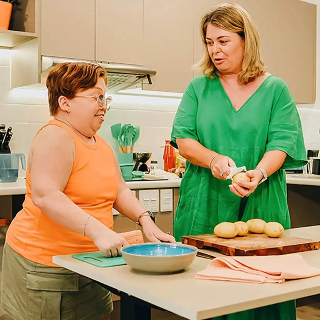 Person with disability prepares food at a low table in their accessible kitchen with support worker.