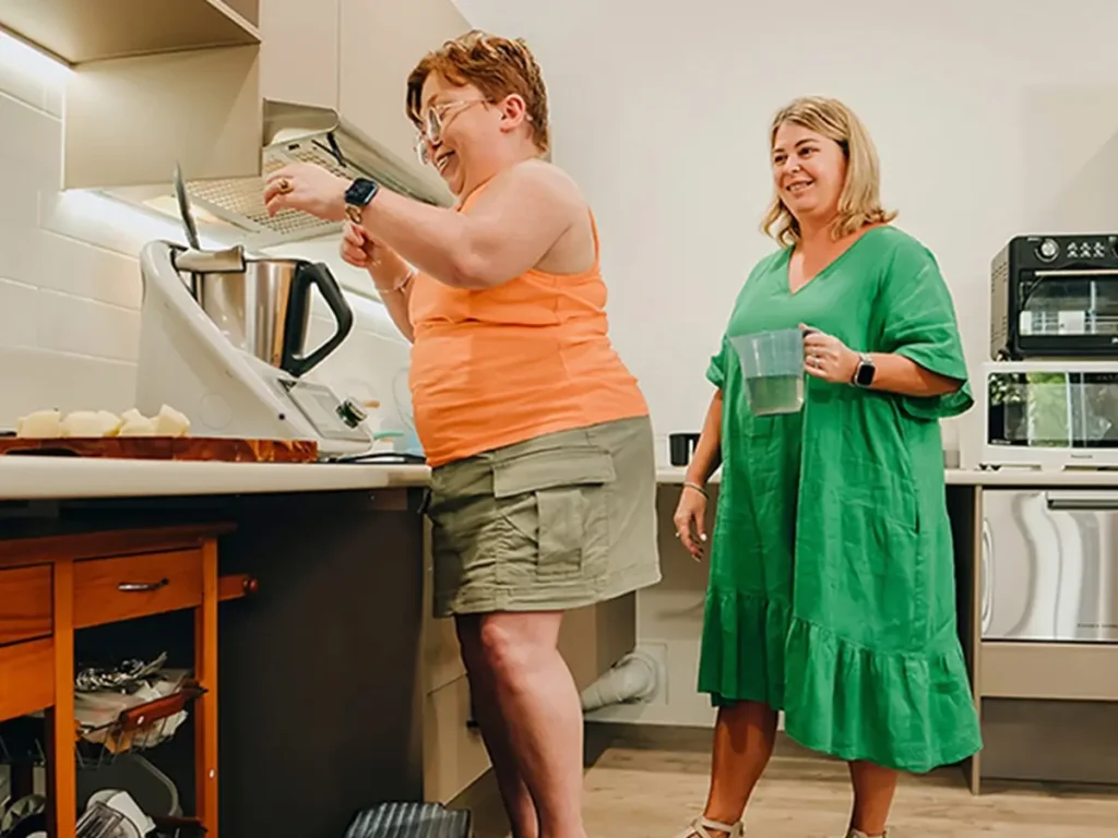 Person with disability stands on step stool at kitchen bench as support worker watches.