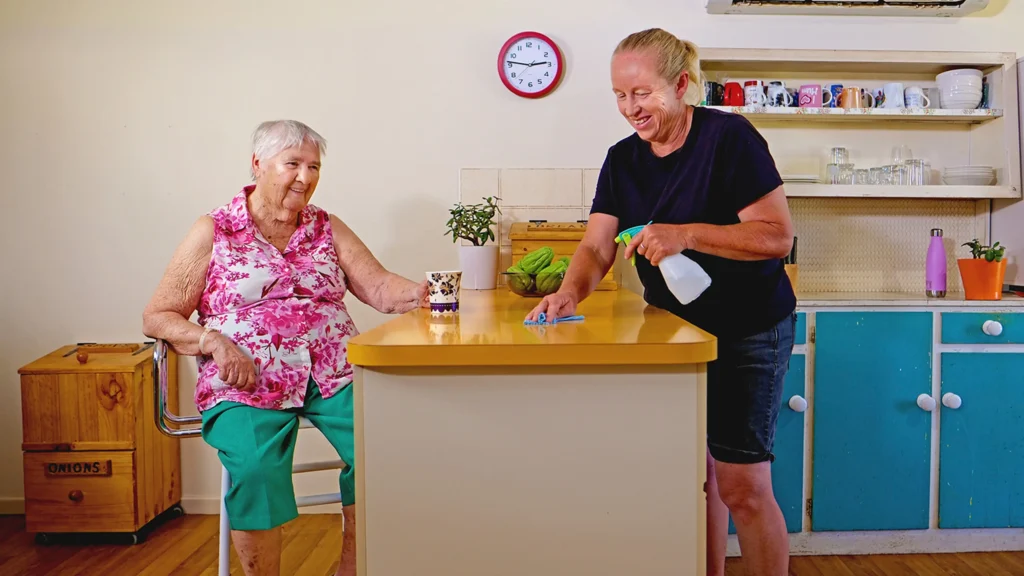 A woman drinks tea while her support worker tidies up.