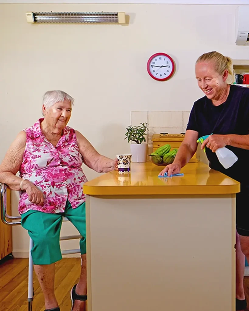 A woman drinks tea while her support worker tidies up.