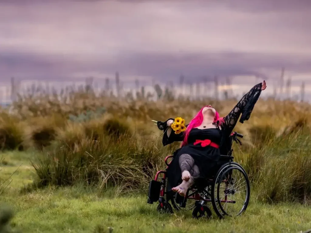 Mobility aid user Zoe Simmons in a field smiling with her arms up