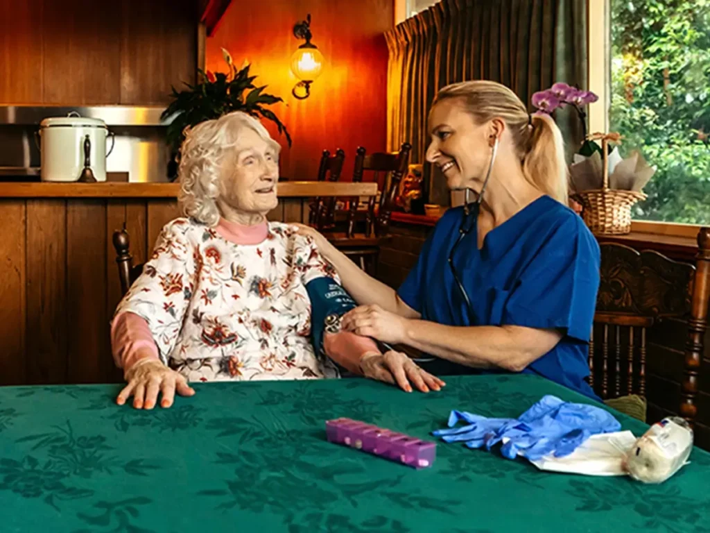 Registered Nurse takes a blood pressure reading for their older client at their dining table. 