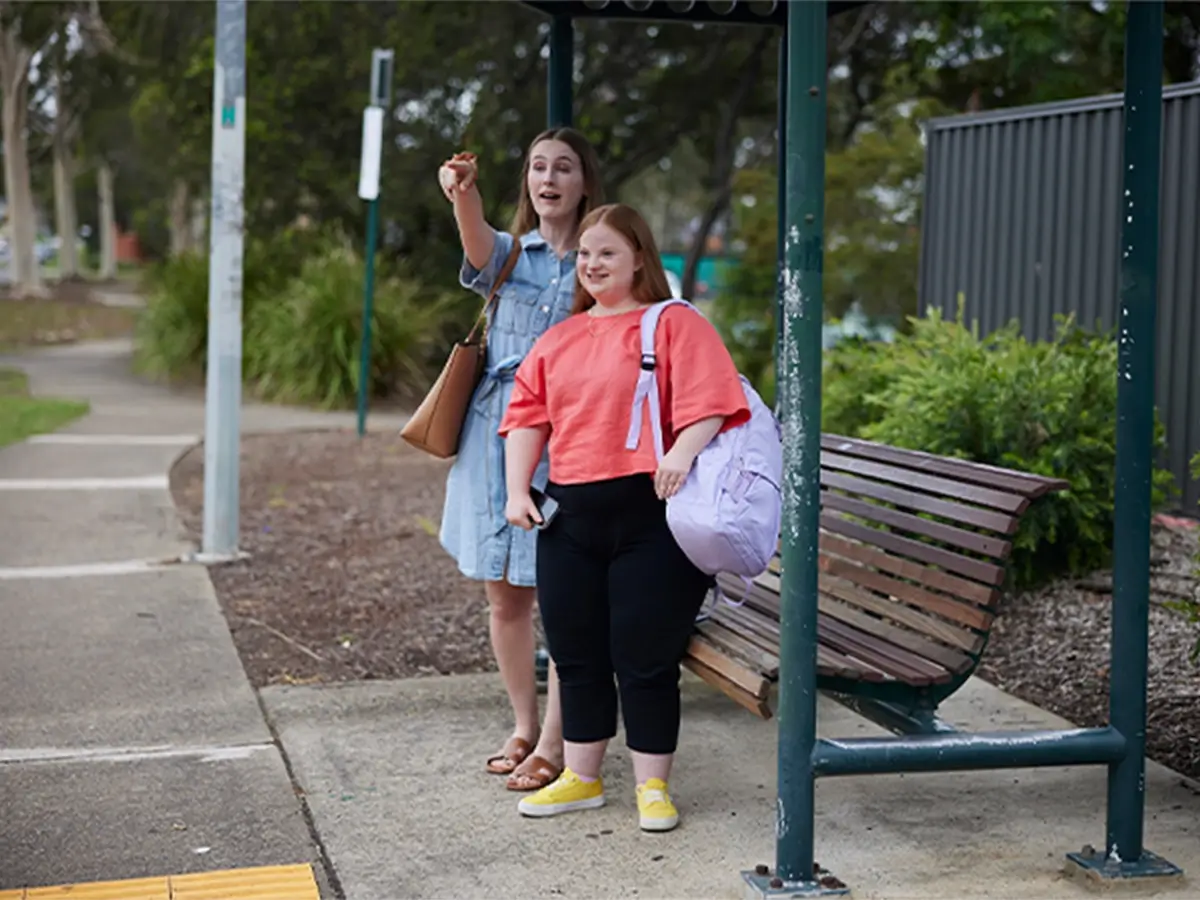 Teenager with Down syndrome standing at bus stop next to support worker who is pointing to a bus.