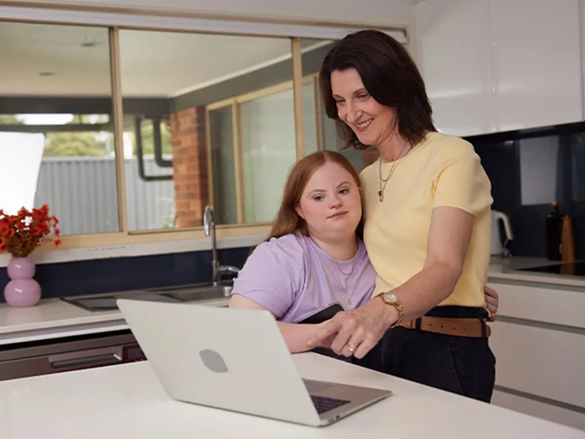 Mum and their teenager with Down syndrome standing in the kitchen while the mum is pointing at a laptop.