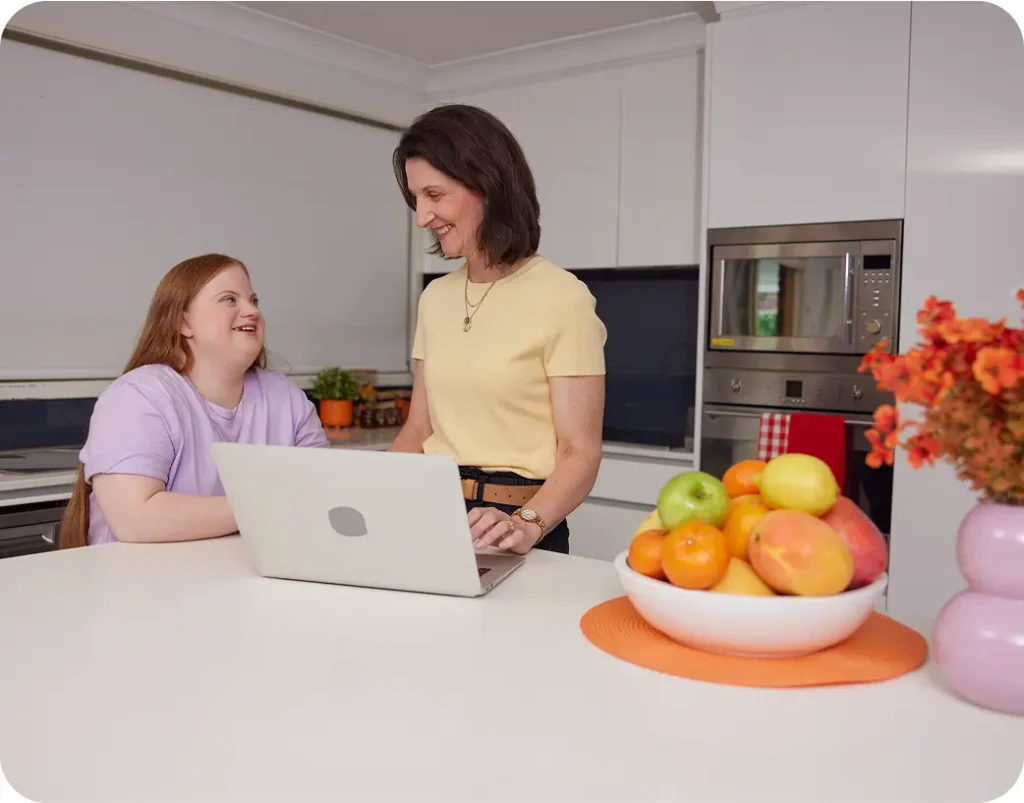 Mum and their teenager with Down syndrome standing in the kitchen looking at a laptop.
