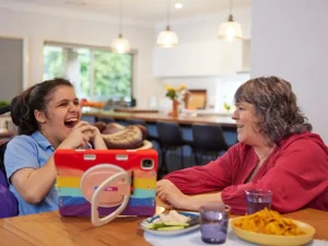 Teenager with Cerebral Palsy and in a wheelchair sitting at a dining table laughing while looking at iPad with support worker.
