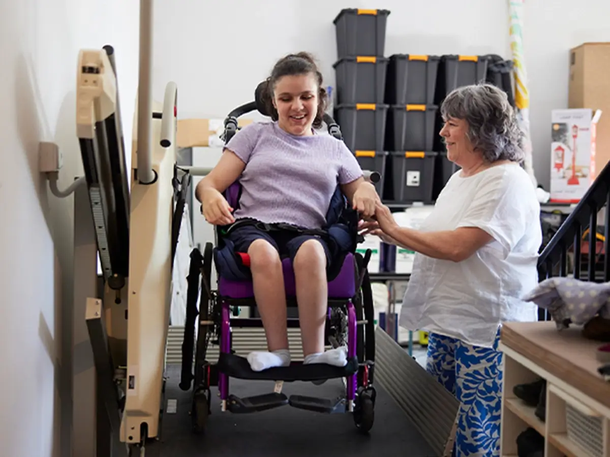 Teenager with Cerebral Palsy and in a wheelchair using platform lift to go up stairs while support worker holds the side of the wheelchair.