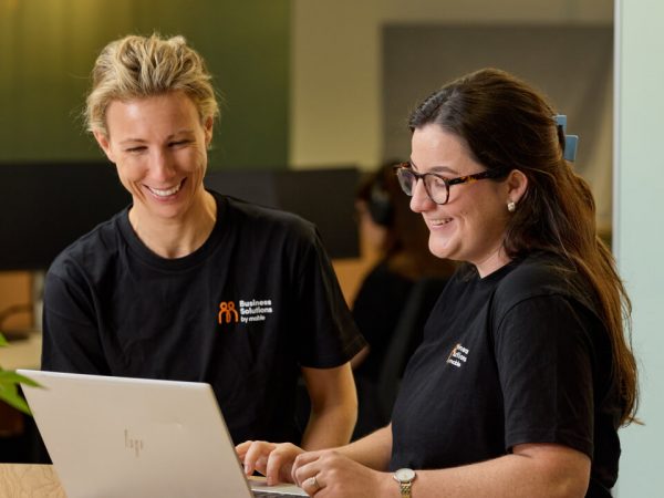 Two smiling women in matching black 'Business Solutions' shirts collaborating and looking at a laptop screen in an office setting.