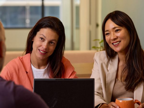 Two smiling professional women, one in an orange blazer and one in a cream blazer, sitting at a meeting table across from an unseen person.