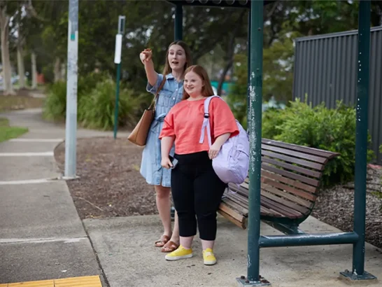 Teenager with Down syndrome standing at bus stop next to support worker who is pointing to a bus.