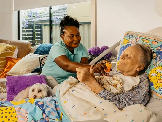 Support worker smiles as they feed an older person living with Dementia laying in bed at home.