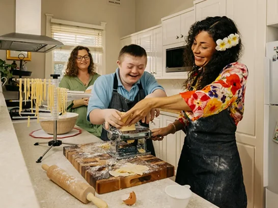 Mum watches support worker and her child with disability roll pasta through dough machine. 