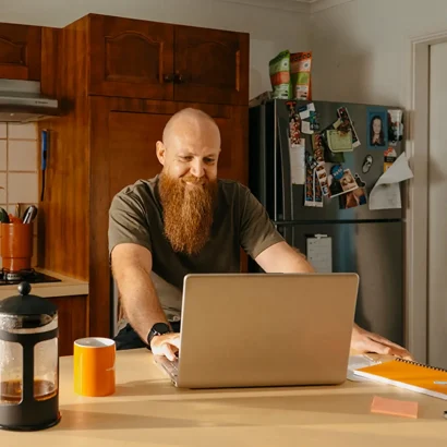 Support worker sits at kitchen bench with laptop and coffee to manage clients on the Mable platform.