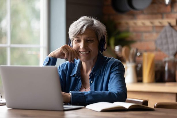 Smiling-older-woman-with-short-grey-hair-wears-headphones-and-works-on-laptop-scaled