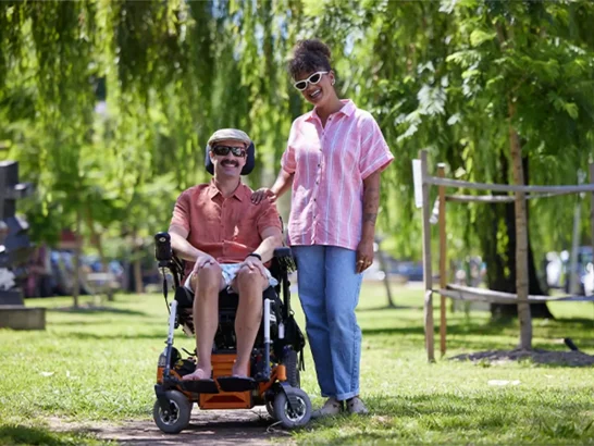 Support worker standing in a park next to client with multiple sclerosis and in wheelchair.