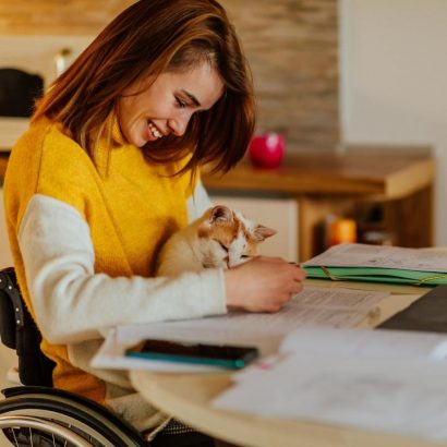A woman in a wheelchair studying at home with her cat in her lap.